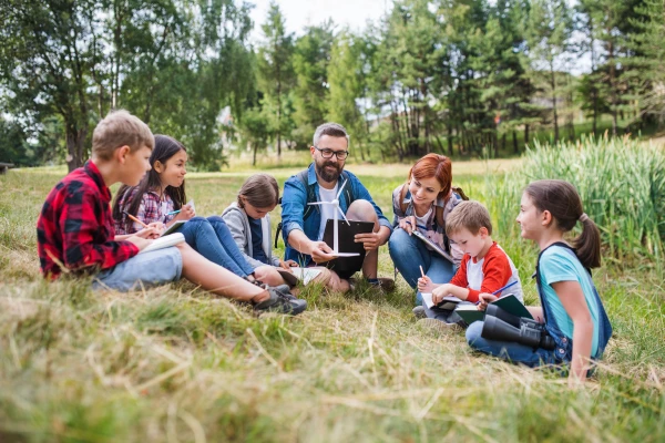 kids sitting in the grass and listening the teacher who hold an model of a wind turbine in hands